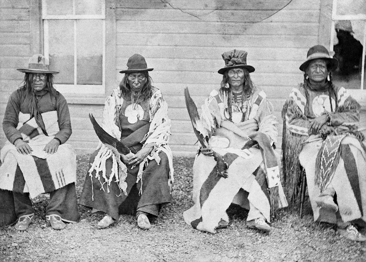 A group of Native Americans sitting in front of a house.