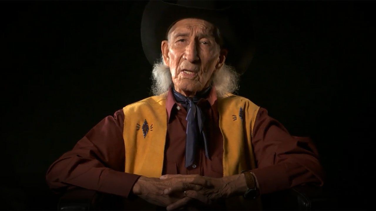 A man in a cowboy hat sits in front of a black background, representing the Head-Smashed-In Buffalo Jump.