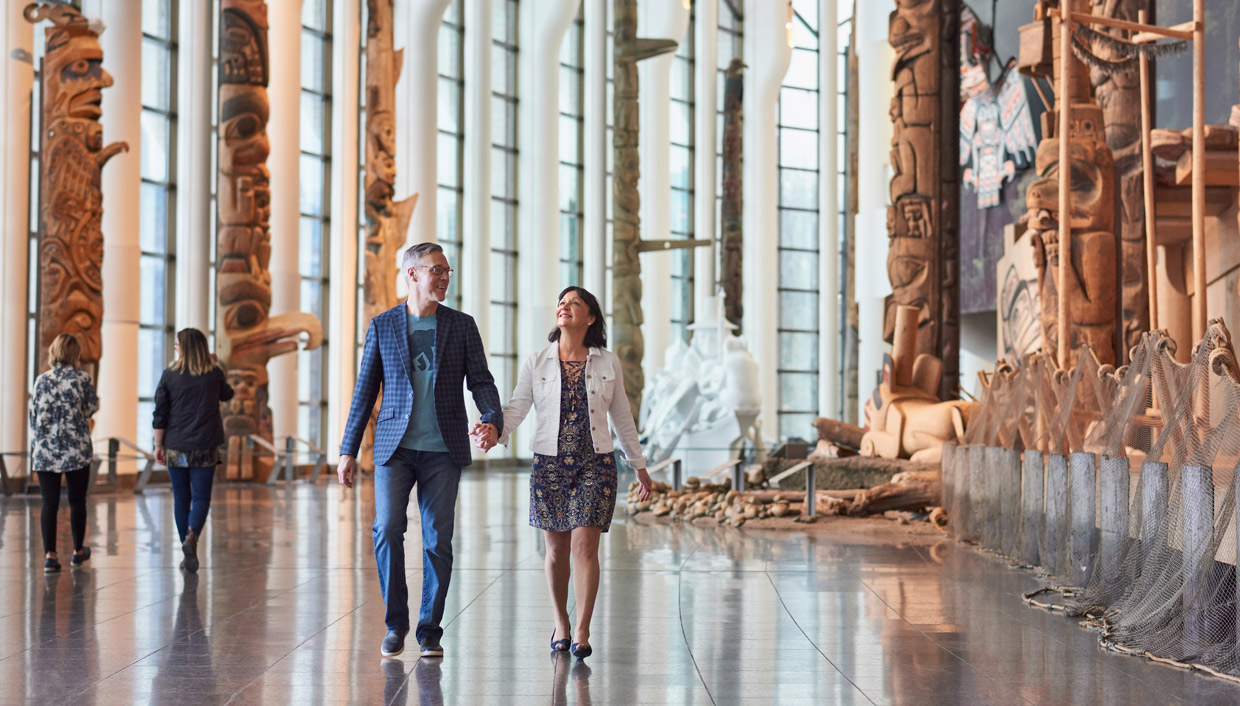 A couple strolling through the Canadian Museum of History in Ottawa, admiring large wooden sculptures.