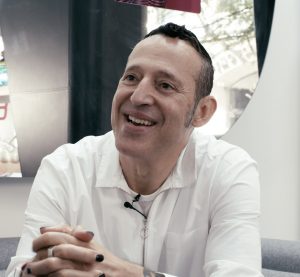 Karim Rashid in a white shirt holds his hands together and smiles.