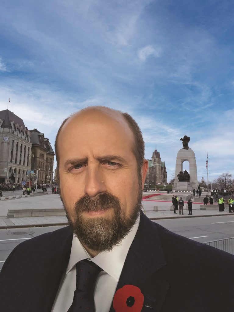 A man in a suit with a poppy on his lapel stands in front of a memorial site in Ottawa.